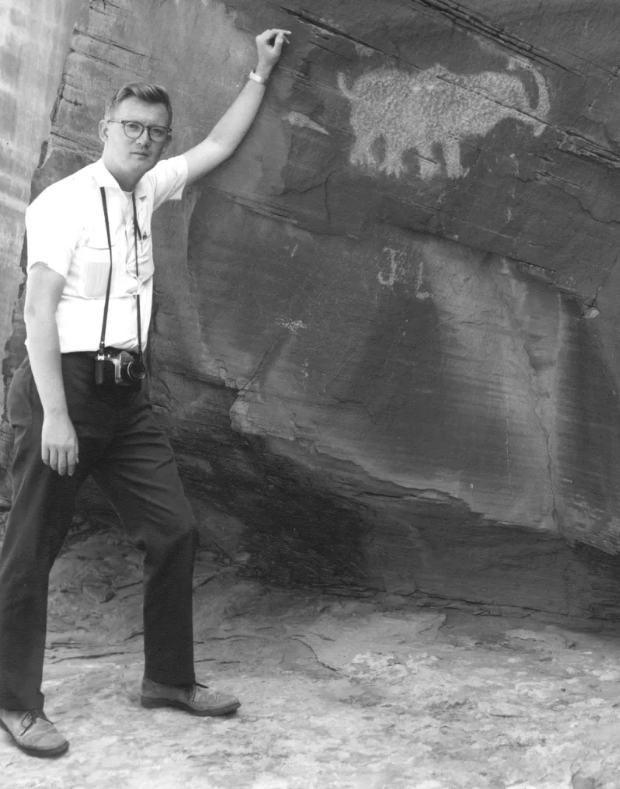 A black and white photo showing a man standing next to a rock boulder; he is leaning against it with one arm up on the rock. On the rock face is a petroglyph that looks like it could be a wooly mammoth.