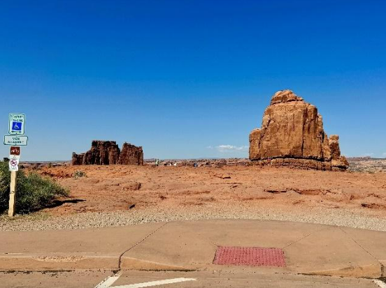 The view and sidewalk at the La Sal Mountain viewpoint: the frame shows an accessible parking spot and sidewalk ramp, plus a view of two sandstone towers.