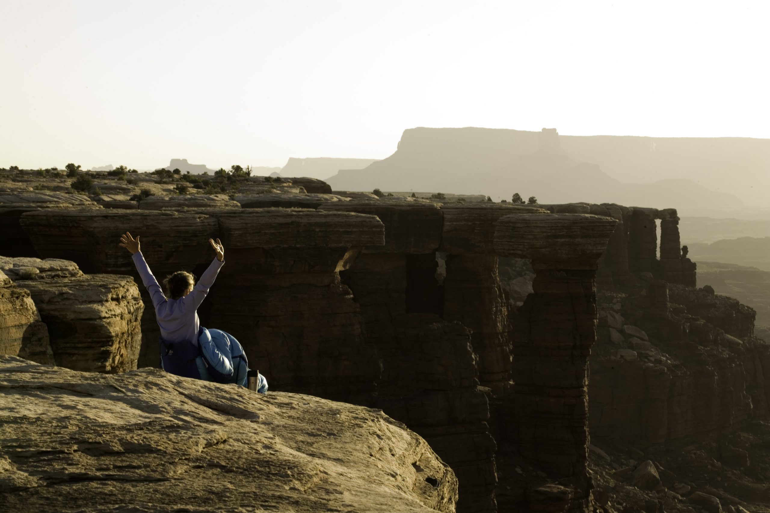 A woman sitting on the edge of a rock overlooking a canyon vista with her arms thrown up into the air.